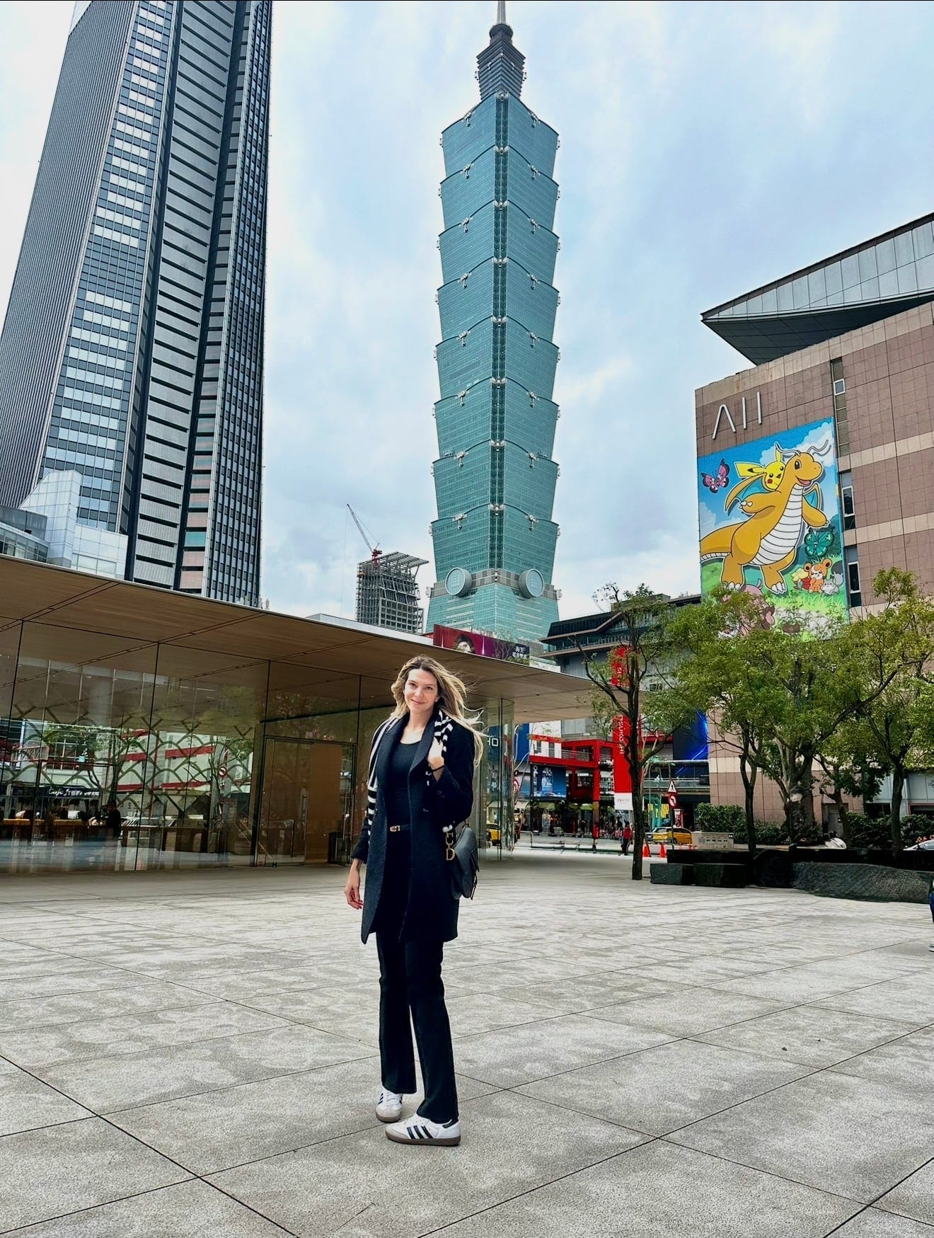 Alexandra in a navy coat in a Taipei plaza with the Taipei 101 tower rising behind her